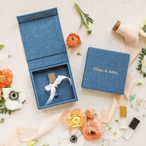 May include: A blue fabric-covered box with a hinged lid, featuring the gold text William & Evelyn on the cover. Inside the open box sits a small metallic object tied with a white ribbon. The set is surrounded by orange and white flowers and ribbons.