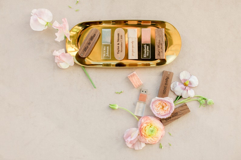May include: A collection of rectangular USB flash drives with wooden and metallic caps, some engraved with names and dates like Taylor & Steven, Elizabeth, and April 2020. The drives are arranged on a gold-toned oval tray and scattered with flowers.