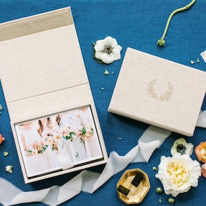 May include: A beige linen-textured keepsake box with a magnetic lid, featuring a gold laurel wreath emblem. One box is open, revealing a photograph of people holding bouquets, while the other is closed. The set is surrounded by various fresh flowers.