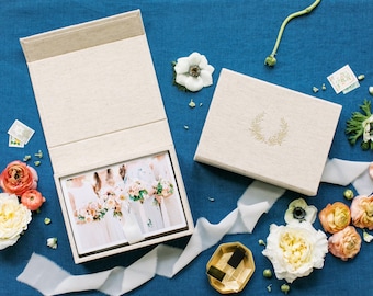 May include: A beige linen-textured keepsake box with a magnetic lid, featuring a gold laurel wreath emblem. One box is open, revealing a photograph of people holding bouquets, while the other is closed. The set is surrounded by various fresh flowers.