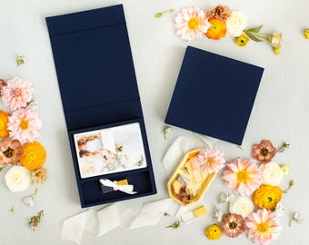 Puede incluir: Dos cajas rectangulares de color azul marino, una abierta y otra cerrada, acompañadas de una pequeña bandeja hexagonal amarilla. La caja abierta contiene una pila de tarjetas rectangulares con imágenes florales y un lazo blanco.