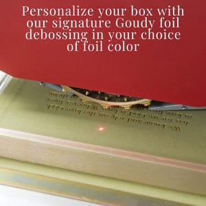 May include: A red foil stamping machine presses text onto a light green surface. The text reads: Personalize your box with our signature Goudy foil debossing in your choice of foil color. Below, the machine imprints: We know and rely on the love God has for us. God is love. Whoever lives in love lives in God.