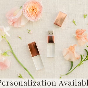 May include: Two clear glass USB drives with rose gold metal tops. The drives are surrounded by pink flowers and greenery. Personalization Available.