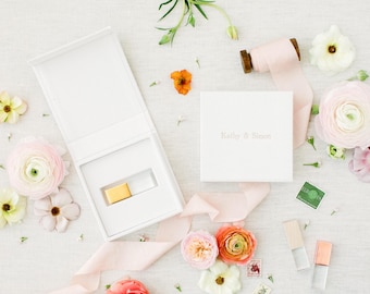 Puede incluir: Un joyero rectangular blanco con tapa abatible, abierto para revelar un pequeño inserto rectangular dorado. Una caja cuadrada blanca cerrada a juego presenta el texto en relieve Kathy & Simon. Una cinta de seda rosa pálido y varias flores rodean las cajas.