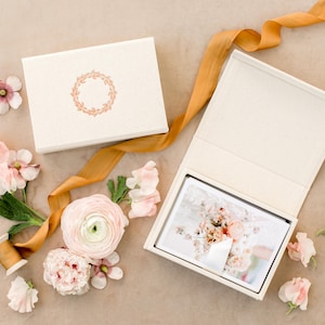 May include: Two off-white photo boxes, one open, with a photograph inside. The closed box has a floral wreath design. A gold ribbon and pink flowers, including sweet peas and ranunculus, are arranged around the boxes.