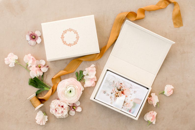 May include: A rectangular off-white box with a gold wreath emblem on the lid, paired with an open matching box containing a photograph of a floral bouquet. A long, flowing mustard-yellow ribbon and several light pink flowers surround the boxes.