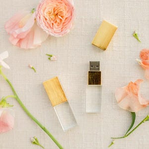 Puede incluir: Dos memorias USB con cuerpo de acrílico transparente y tapas de madera clara, dispuestas junto a varias flores de color rosa suave y melocotón. Una memoria se muestra sin tapa, revelando el conector metálico, mientras que la otra está cerrada.
