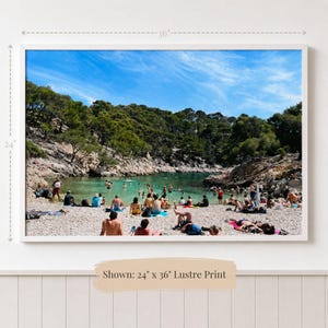 May include: A landscape photograph of a pebble beach and turquoise cove filled with swimmers under a bright blue sky. The image is framed in white and measures 24 inches by 36 inches, featuring a dense forest on the rocky cliffs surrounding the water.