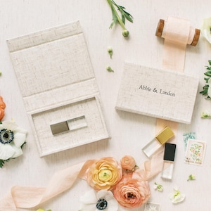 Op de afbeelding: Een beige linnen-textuur doosset voor een USB-stick, met de tekst Abbie & Landon op het deksel. De set bevat een open doos met een zilveren USB-stick, een gesloten doos, een perzikkleurig lint en diverse oranje en witte bloemen.