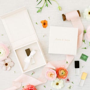 May include: An overhead shot of wedding-themed items. A white box with a ribbon and a wooden item inside is open. A smaller box with "Annie & James" is next to it. Pink and orange flowers and ribbon are scattered around.