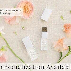 K&ouml;nnte beinhalten: Zwei wei&szlig;e USB-Sticks mit abnehmbaren Kappen und individuell graviertem Text, einschlie&szlig;lich Namen und Daten. Die Sticks sind von pfirsich- und rosafarbenen Blumen umgeben, mit dem sichtbaren Text Add names, dates, branding, or a special message.