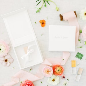 May include: A white square gift box with a hinged lid, featuring the embossed text "Annie & James" on the cover. The box is accompanied by a spool of light pink silk ribbon, two small glass vials with caps, and various fresh ranunculus flowers.