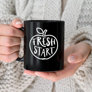 Op de afbeelding: Zwarte koffiemok met een witte appel afbeelding en de tekst "Fresh Start".