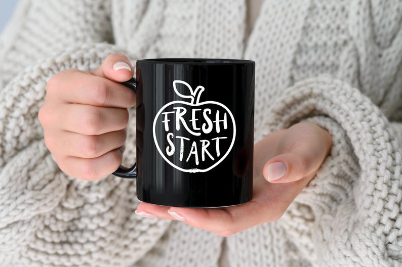 Op de afbeelding: Zwarte koffiemok met een witte appel afbeelding en de tekst "Fresh Start".