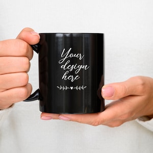 May include: A black ceramic mug with a white handle. The mug has the text "Your design here" written on it in a white cursive font.