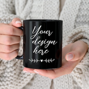 Op de afbeelding: Een zwarte keramische mok met een witte handgreep. De mok heeft de tekst "Your design here" erop geschreven in een cursief schrift. Aan weerszijden van de tekst staan kleine witte bloemmotieven.