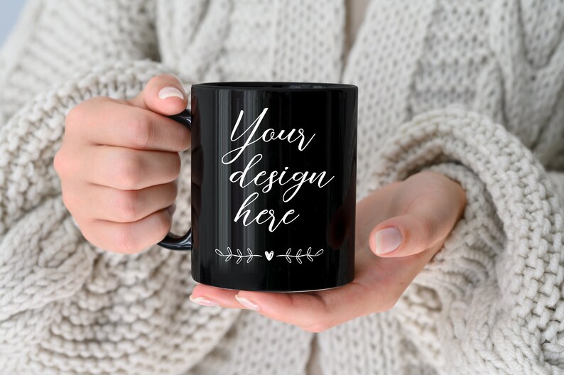 Op de afbeelding: Een zwarte keramische mok met een witte handgreep. De mok heeft de tekst "Your design here" erop geschreven in een cursief schrift. Aan weerszijden van de tekst staan kleine witte bloemmotieven.