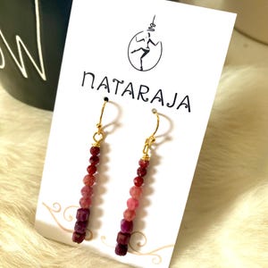 May include: Dangle earrings with gold-coloured hooks and a gradient of red and pink beads. The earrings are displayed on a white card with the word "NATARAJA" and a graphic of a dancing figure.