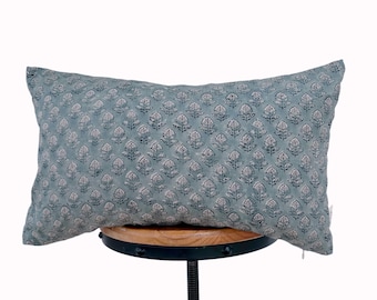 May include: A rectangular throw cushion in a muted sage green colour featuring a repeating dark blue floral block print pattern. The cushion has a visible white zip pull on the right side and is displayed on a round wooden and black metal stool.