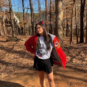 May include: A woman wearing a red jacket with a white t-shirt that says "Who's Travis Once Away?" and a black skirt. She is standing in a wooded area.