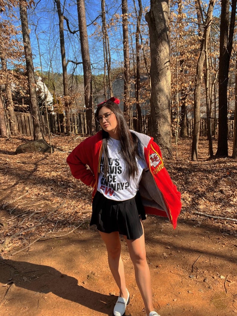 May include: A woman wearing a red jacket with a white t-shirt that says "Who's Travis Once Away?" and a black skirt. She is standing in a wooded area.