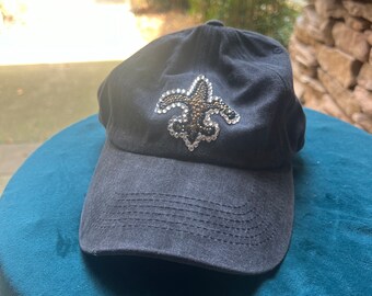 May include: A black baseball cap with a curved brim. The cap features a jeweled fleur-de-lis emblem on the front. The cap is sitting on a teal surface.