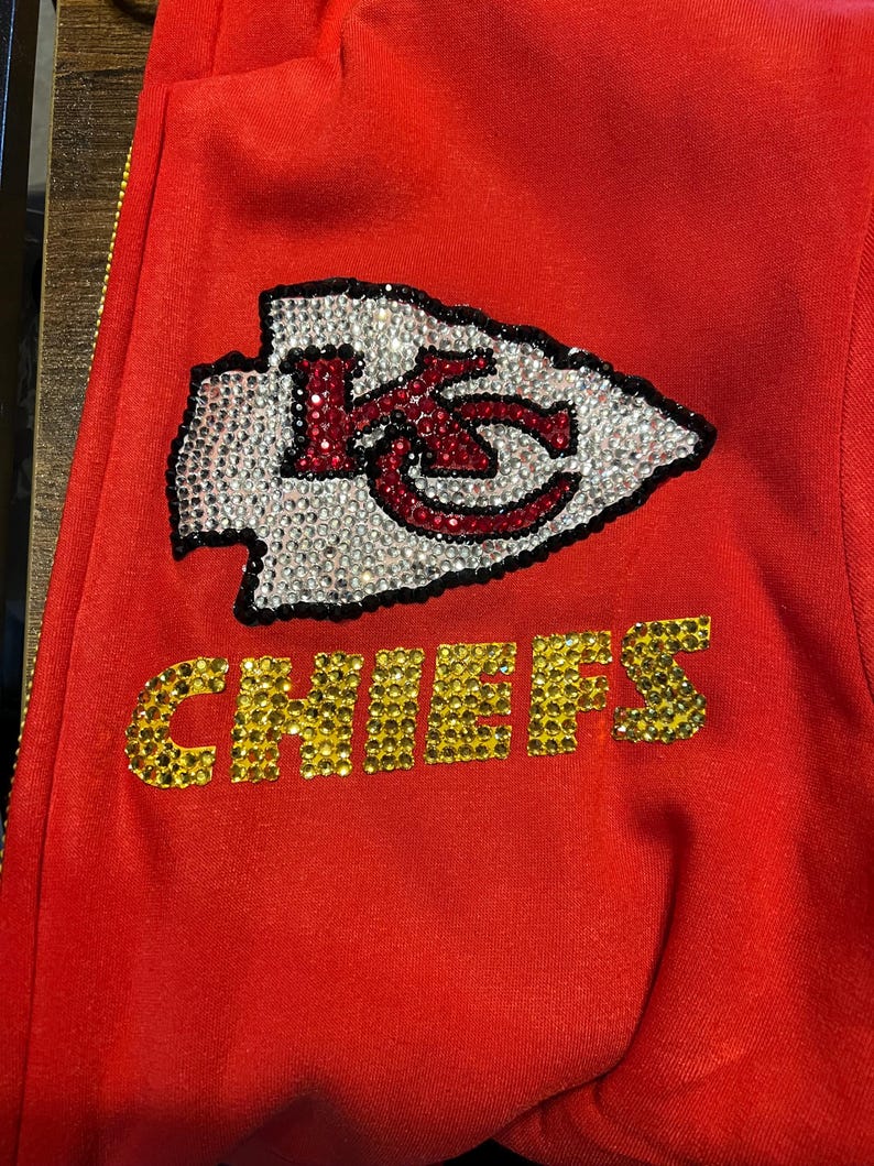 May include: Red sweatshirt with a rhinestone Kansas City Chiefs logo and the word "CHIEFS" in rhinestones.