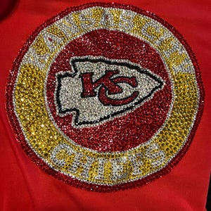 May include: A red, white, and gold rhinestone design of the Kansas City Chiefs logo. The logo is surrounded by a circle of rhinestones that spell out "Kansas City Chiefs".