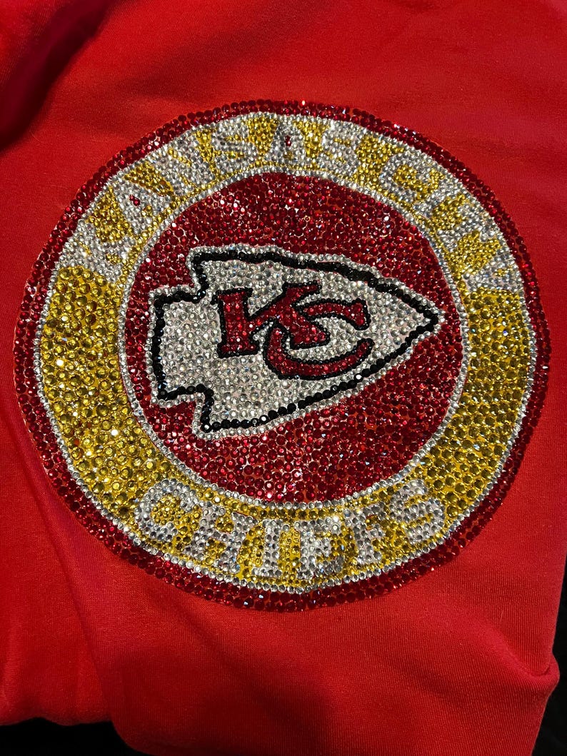May include: A red, white, and gold rhinestone design of the Kansas City Chiefs logo. The logo is surrounded by a circle of rhinestones that spell out "Kansas City Chiefs".