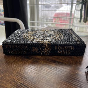 May include: A black book with gold lettering that reads "Rebecca Yarros" and "Fourth Wing". The book is covered in rhinestones.