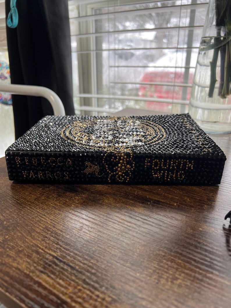 May include: A black book with gold lettering that reads "Rebecca Yarros" and "Fourth Wing". The book is covered in rhinestones.