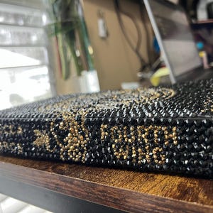 May include: A black book covered in black and gold rhinestones. The book is lying on a wooden surface.