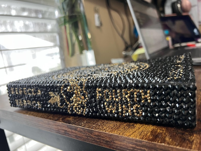 May include: A black book covered in black and gold rhinestones. The book is lying on a wooden surface.