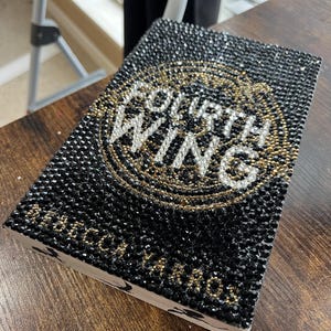 May include: A black book covered in black and gold rhinestones with the words "Fourth Wing" in white lettering.