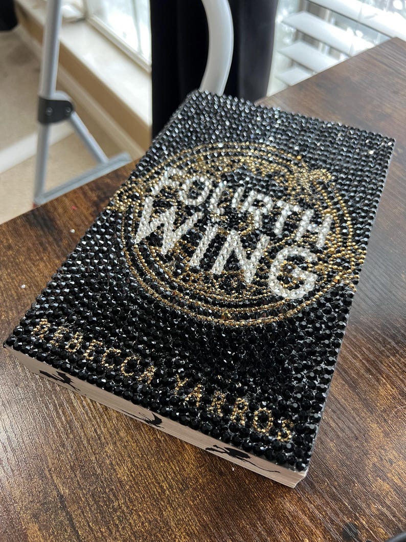 May include: A black book covered in black and gold rhinestones with the words "Fourth Wing" in white lettering.