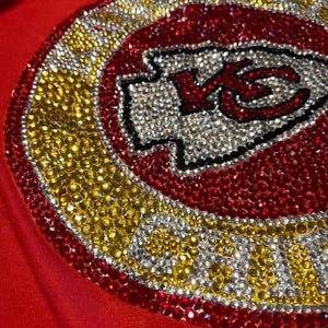 May include: A red shirt with a large, rhinestone-encrusted Kansas City Chiefs logo. The logo is surrounded by a gold circle with the words "Chiefs Kingdom" in rhinestones.