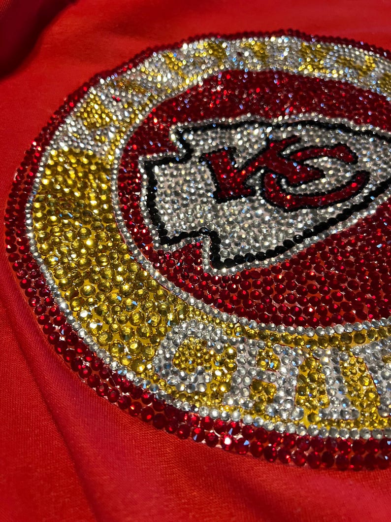 May include: A red shirt with a large, rhinestone-encrusted Kansas City Chiefs logo. The logo is surrounded by a gold circle with the words "Chiefs Kingdom" in rhinestones.