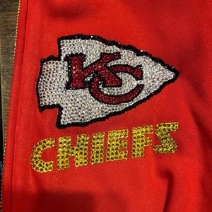 May include: Red sweatshirt with a large rhinestone Kansas City Chiefs logo. The logo is a red and white arrowhead with the letters "KC" inside. The word "CHIEFS" is below the arrowhead in gold rhinestones.