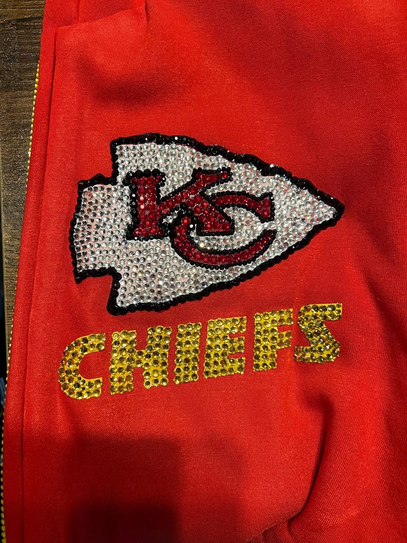 May include: Red sweatshirt with a large rhinestone Kansas City Chiefs logo. The logo is a red and white arrowhead with the letters "KC" inside. The word "CHIEFS" is below the arrowhead in gold rhinestones.