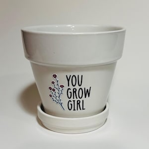 以下が含まれることがあります： 白いセラミック製のプランターと受け皿。プランターには、「You Grow Girl」と小さな花のデザインが書かれた黒と白のステッカーが貼られています。