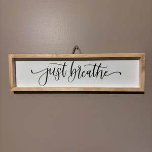 May include: A wooden framed sign with the text "just breathe" in black cursive lettering on a white background.