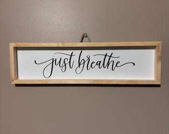 Puede incluir: Un letrero enmarcado de madera con el texto "just breathe" en letras cursivas negras sobre un fondo blanco.