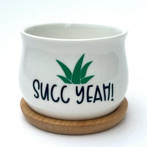 Puede incluir: Una maceta de cerámica blanca con silueta curva, que presenta un gráfico verde de una planta suculenta y el texto negro SUCC YEAH! impreso en el frente. La maceta descansa sobre una base circular de platillo de madera de bambú clara.