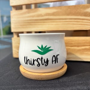May include: A white ceramic planter with a rounded shape, featuring a green succulent graphic and the black text "thirsty AF" printed on the front. The planter sits on a circular light brown bamboo wood base, providing a stable foundation.
