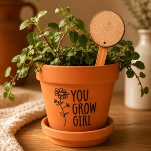 Puede incluir: Una maceta de terracota con su plato a juego que contiene una planta verde. La maceta presenta un gráfico negro de una flor y el texto "YOU GROW GIRL" impreso en el frente. Un marcador de plantas de madera redondo está insertado en la tierra.