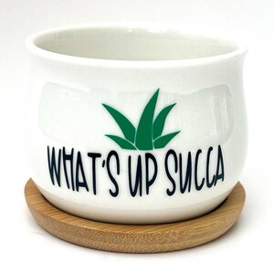 Puede incluir: Una maceta de cerámica blanca de forma redondeada, con un gráfico verde de una planta suculenta y el texto negro "WHAT'S UP SUCCA" impreso en el frente. La maceta descansa sobre una bandeja base circular de madera de color claro.