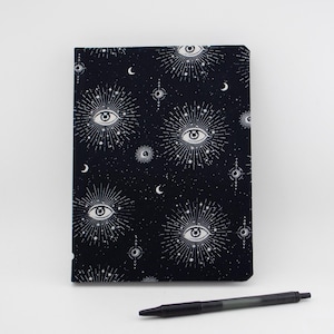 May include: A rectangular notebook with a black cover featuring a white repeating pattern of eyes surrounded by radiating lines, stars, and crescent moons. A black retractable ballpoint pen is positioned horizontally below the notebook.