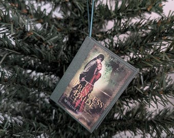 May include: A miniature book ornament featuring a cover illustration of two figures from The Princess Bride by William Goldman. The book has a dark gray spine and edges, and it hangs from a thin teal ribbon, displaying the title and author name.
