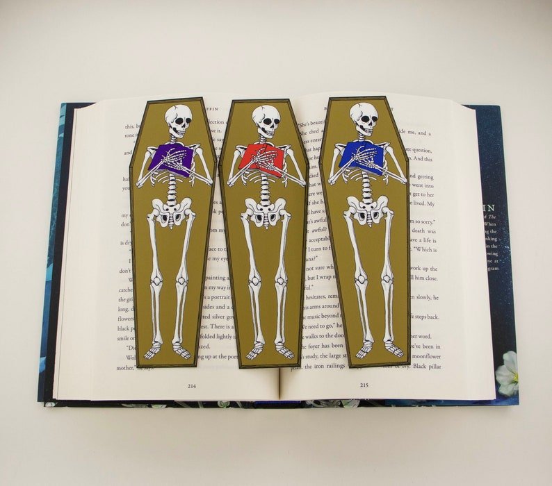May include: Three gold coffin-shaped bookmarks featuring skeletons holding books. The skeletons are wearing purple, red, and blue shirts. The bookmarks are numbered 214, 215, and 216.