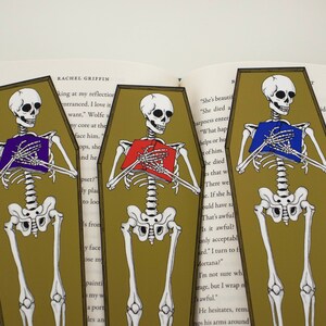 May include: Three skeleton bookmarks with different coloured books in their hands. The bookmarks are shaped like coffins and have a gold background. The text on the bookmarks reads "She's beautiful", "She died a", "harpness enter", "What hap", "helps or hu", "t of him", "he died", "es that w", "There we", "e. If she h", "ould have sa", "That's awful", "Is it awful?", "only acceptabl", "ked." I turn to f", "Mortana?", "I'm not sure wh", "arage, but I wrap n", "sawful.", "He hesitates, rem", "raps his arms around", "ide me", "ate o", "nd", "W", "m", "d", "av".